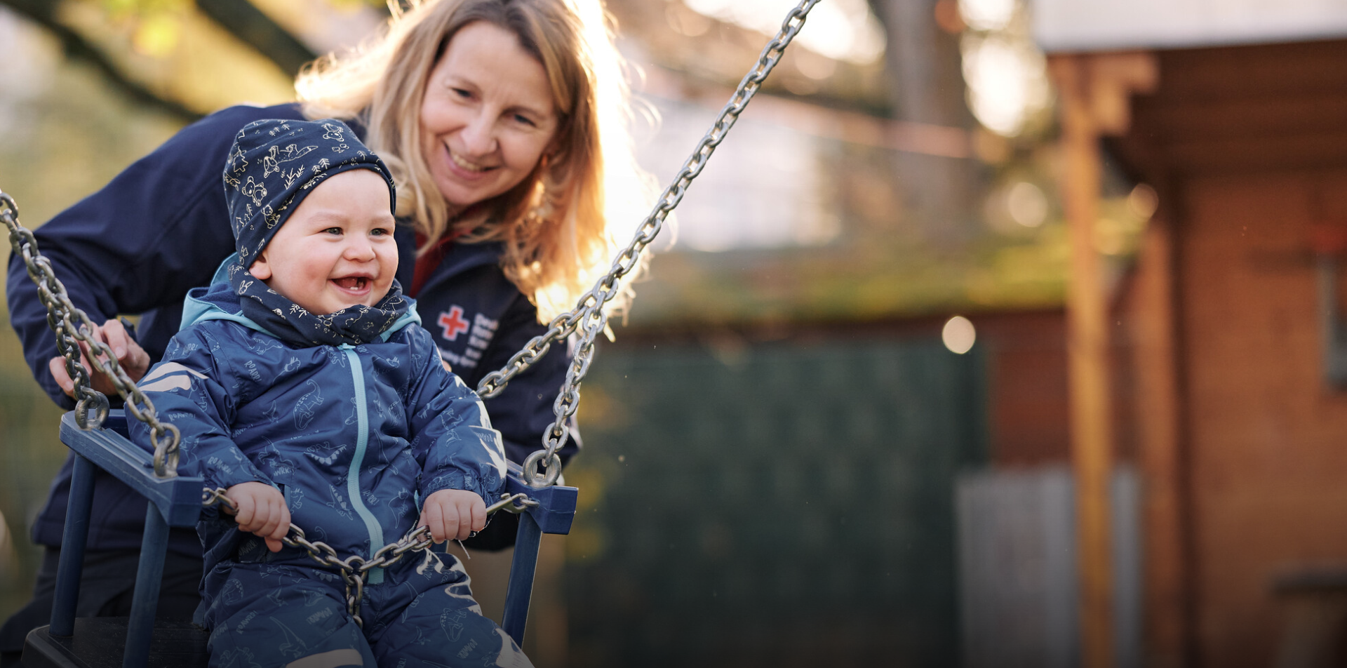 Eine Erzieherin steht hinter einem Kleinkind, das gerade im Garten schaukelt.