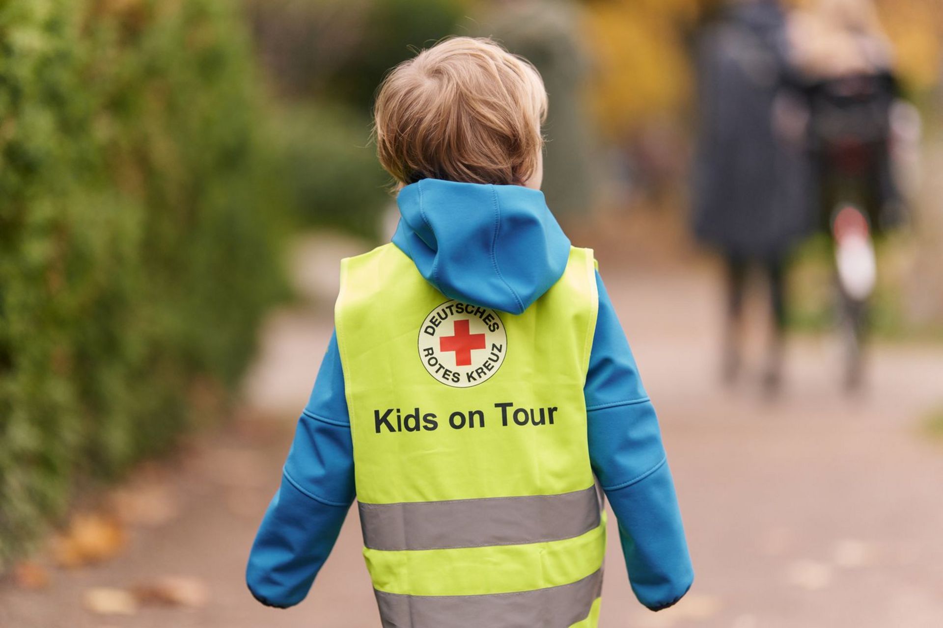 Ein kleines Kind trägt eine Leuchtweste für Kita-Kinder mit der Aufschrift ‚Kids on Tour‘ und dem DRK-Logo.
